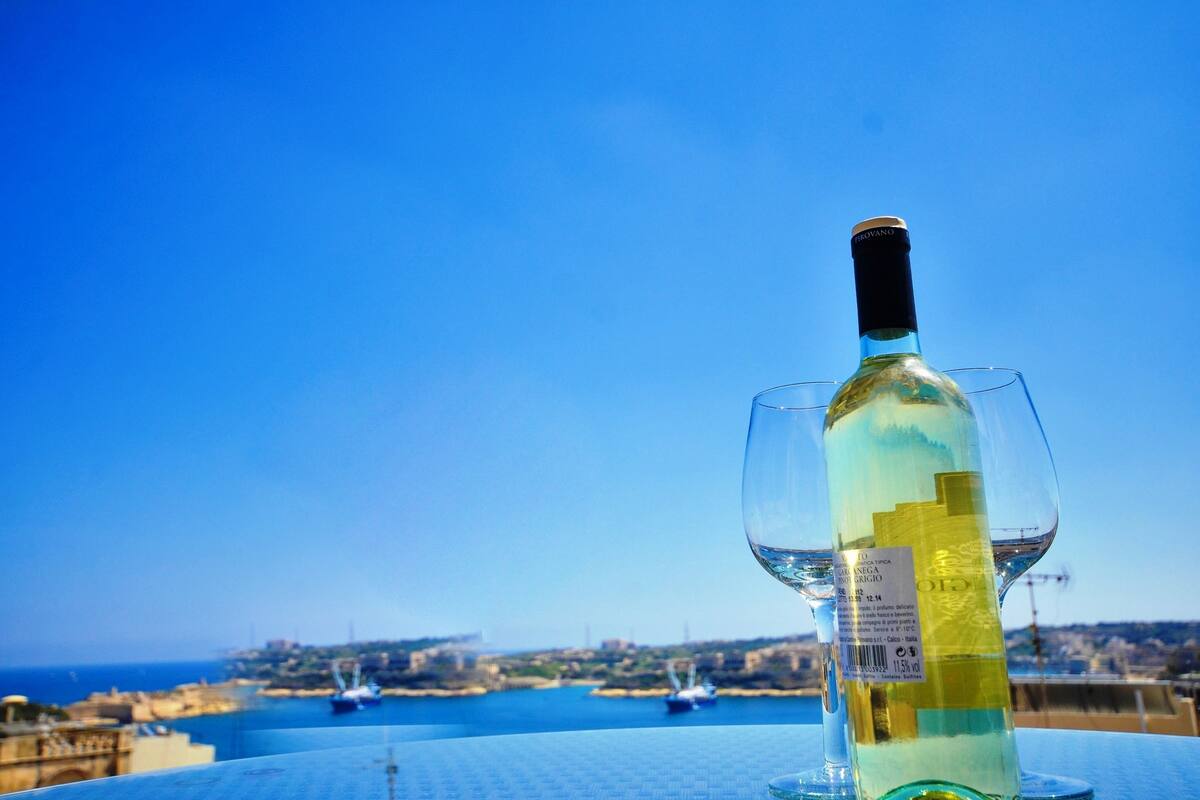 Una botella de vino blanco y una copa sobre una mesa, con vista a un pueblo costero y barcos al fondo.
