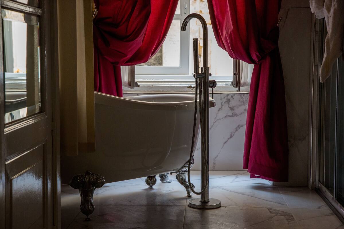 Un baño con una bañera independiente, un ventanal con cortinas rojas y suelo de mármol.