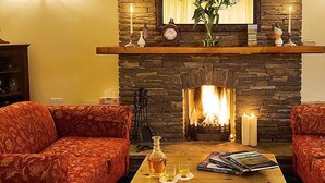 A cozy living room with a stone fireplace, two patterned sofas, a coffee table with books and a bottle, and a clock on the mantel. - Lehinch Lodge (Lahinch)