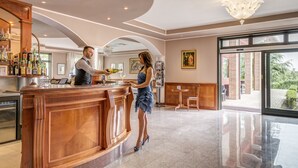 A bar area with a wooden counter, a bartender, and a customer. There are framed pictures on the wall, a chandelier, and a large window with a view of greenery. - Hotel SantaMaria Langhe (La Morra)
