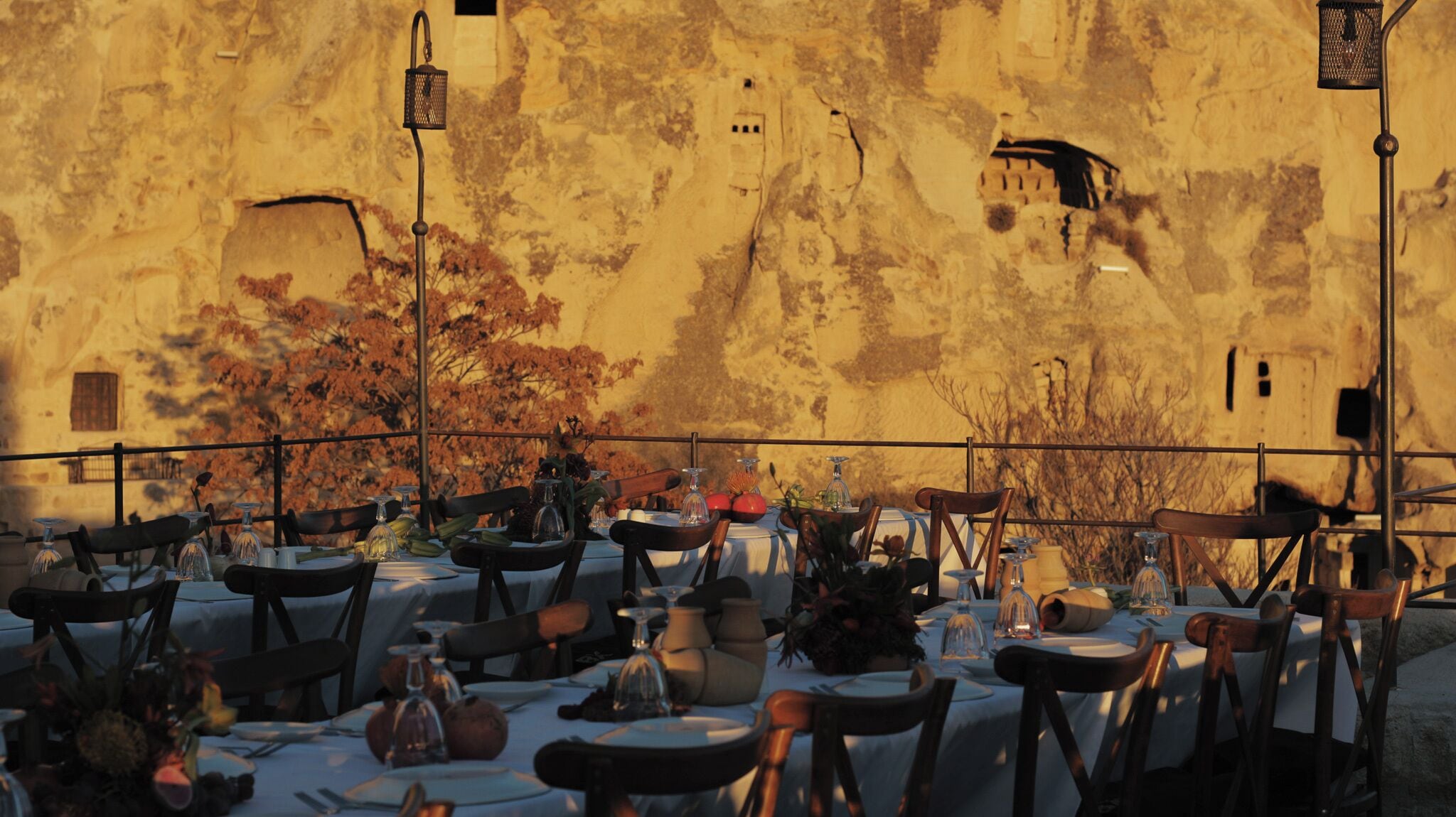 Area pranzo all'aperto con tavoli apparecchiati per due, con a sfondo un muro in pietra rustica e una ringhiera in metallo.
