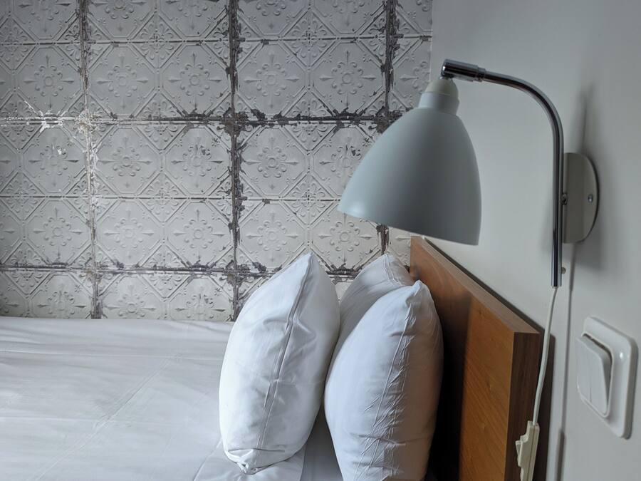 A neatly made bed with white linens and two pillows, a modern wall-mounted lamp, and a patterned tile backsplash.