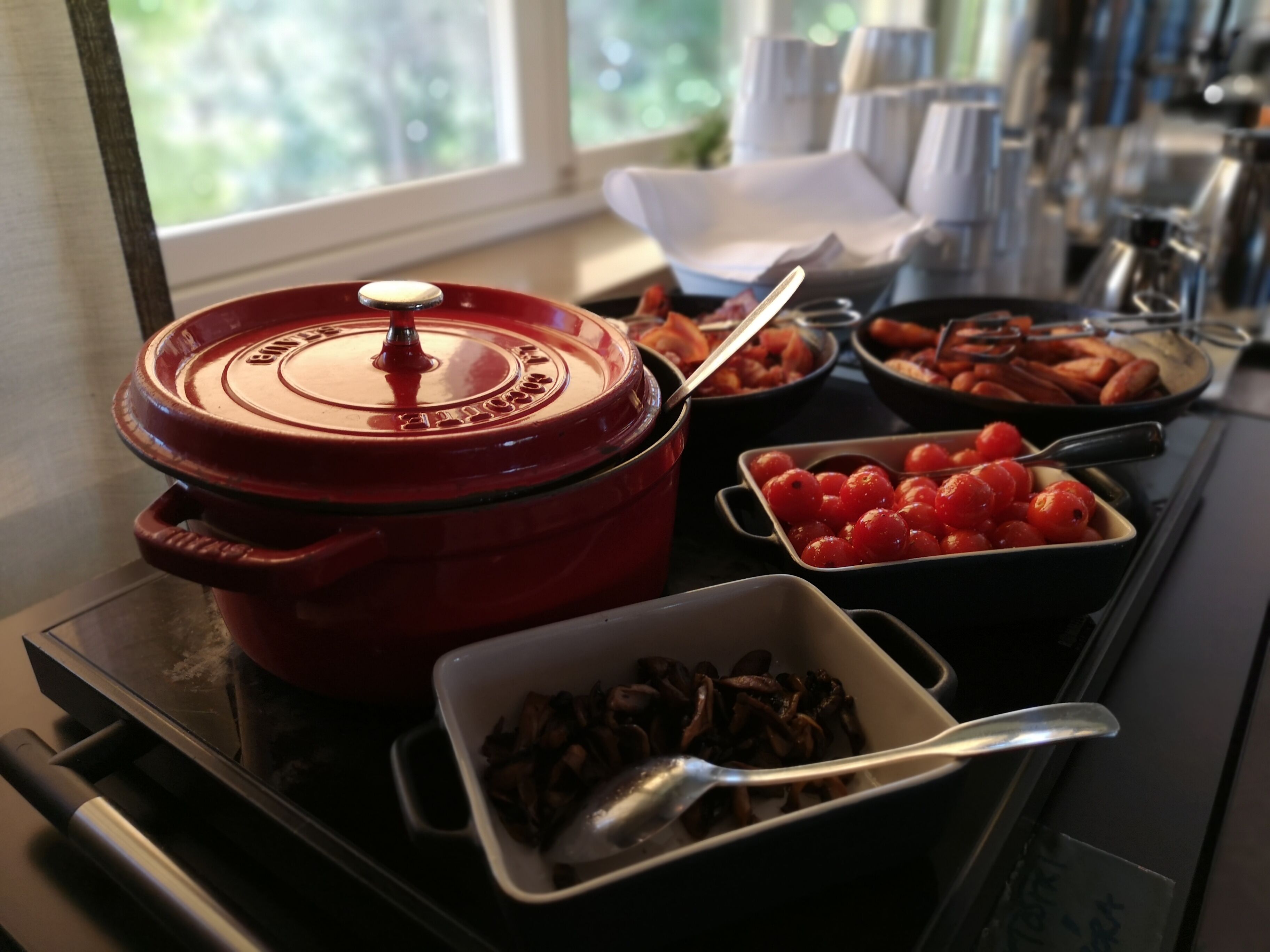 Ein reichhaltiges Buffet mit verschiedenen Speisen, darunter ein roter Topf, Schalen mit Kirschtomaten und eine Schale mit Pilzen.