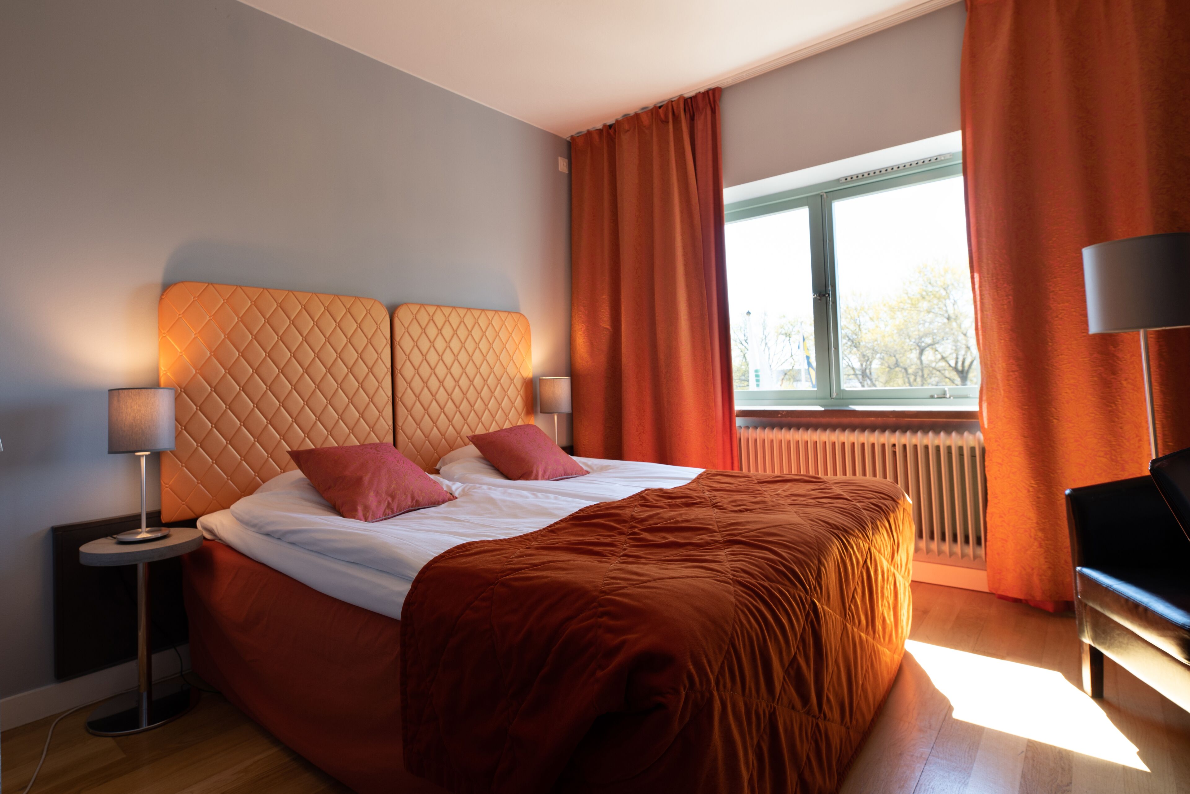 A modern hotel room with a large bed, two bedside lamps, a view of the outdoors through a window, and a radiator.