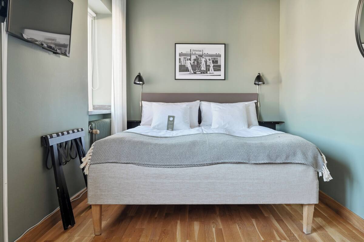 A neatly arranged bedroom with a bed, bedside lamps, a framed picture on the wall, and a radiator.