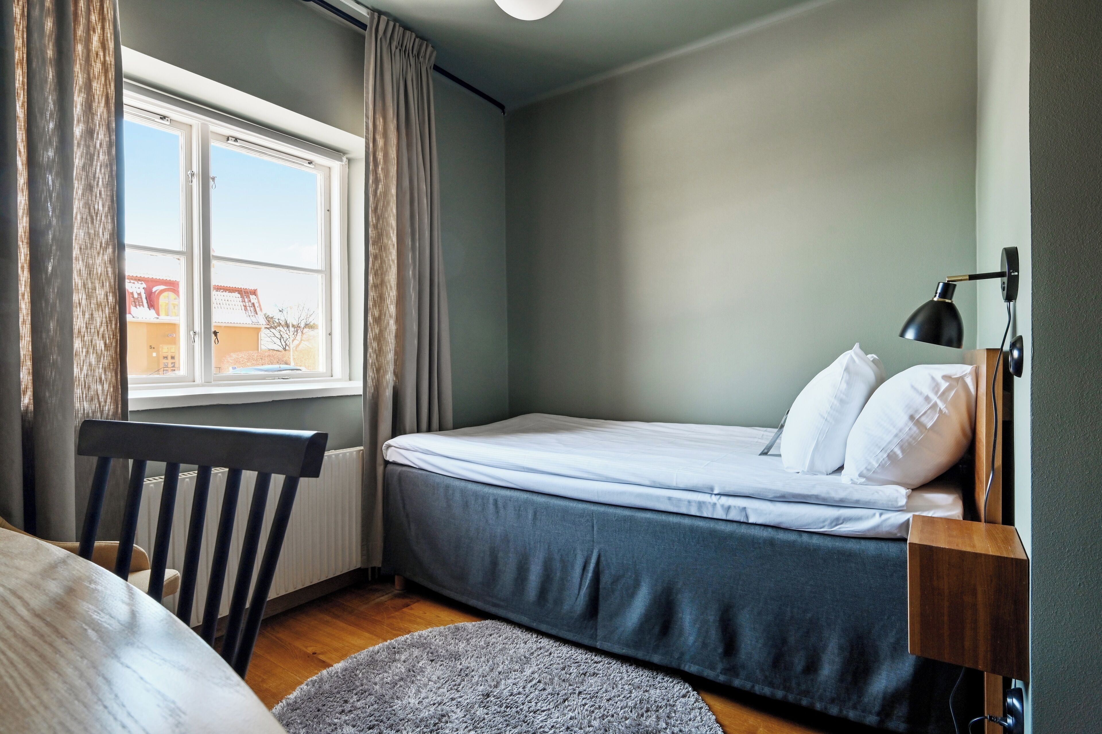 A bedroom with a bed, a chair, a window with curtains, a lamp, and a radiator.