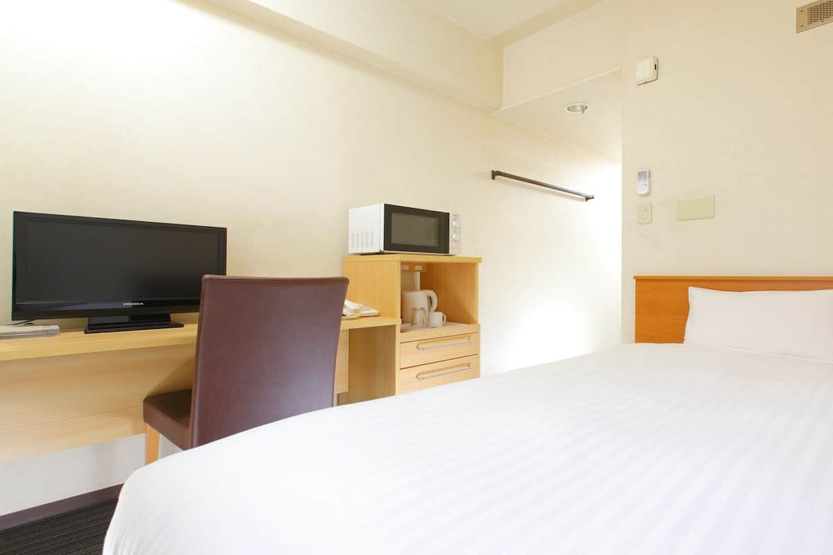Quarto de hotel com cama, escrivaninha, televisão, micro-ondas e uma cadeira.
