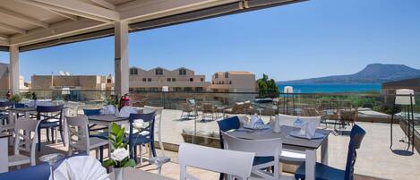 An outdoor dining area with tables and chairs, a clear view of the sea and buildings in the background.