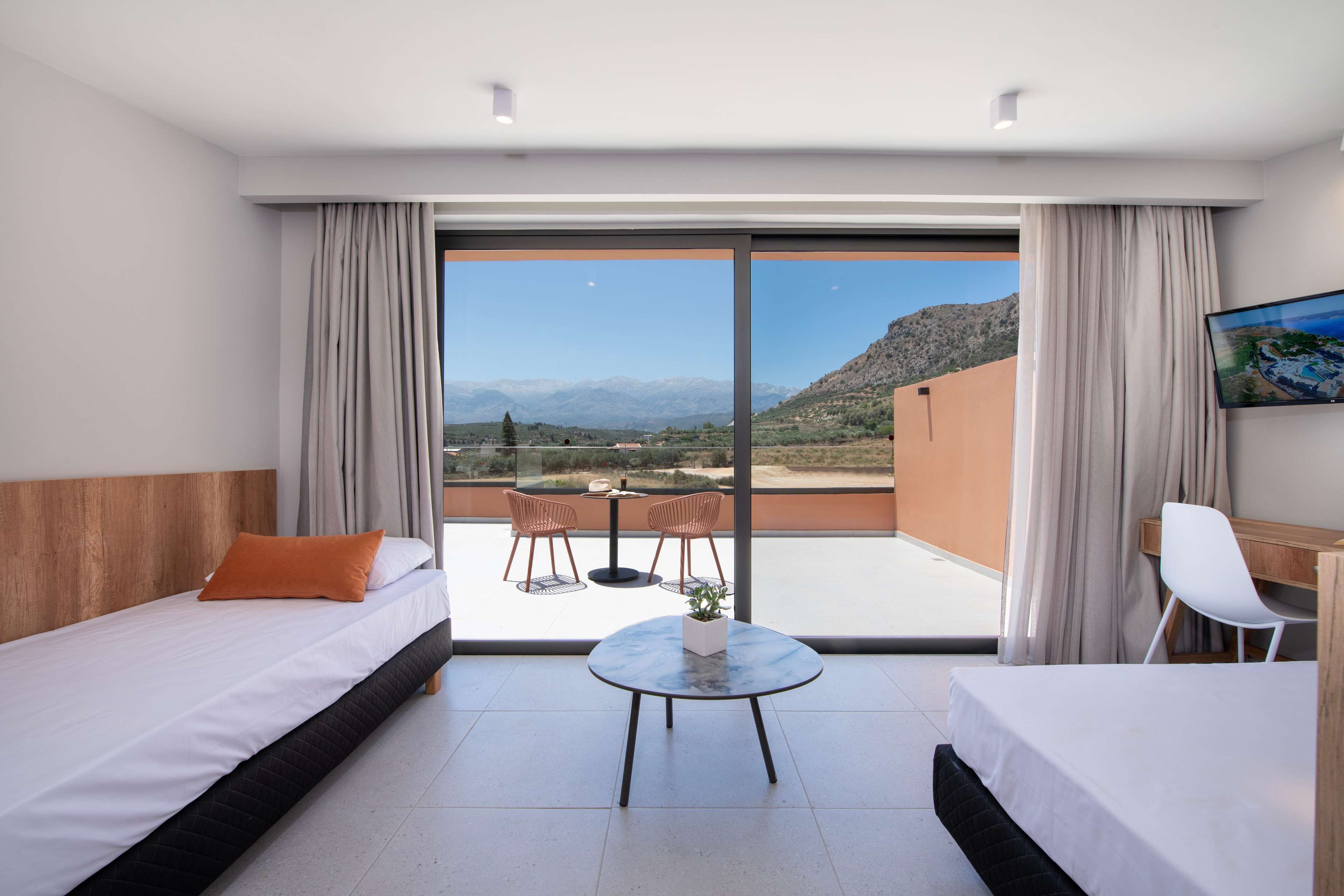 A hotel room with two beds, a small table, and a chair. There is a balcony with a view of mountains and a table with chairs.