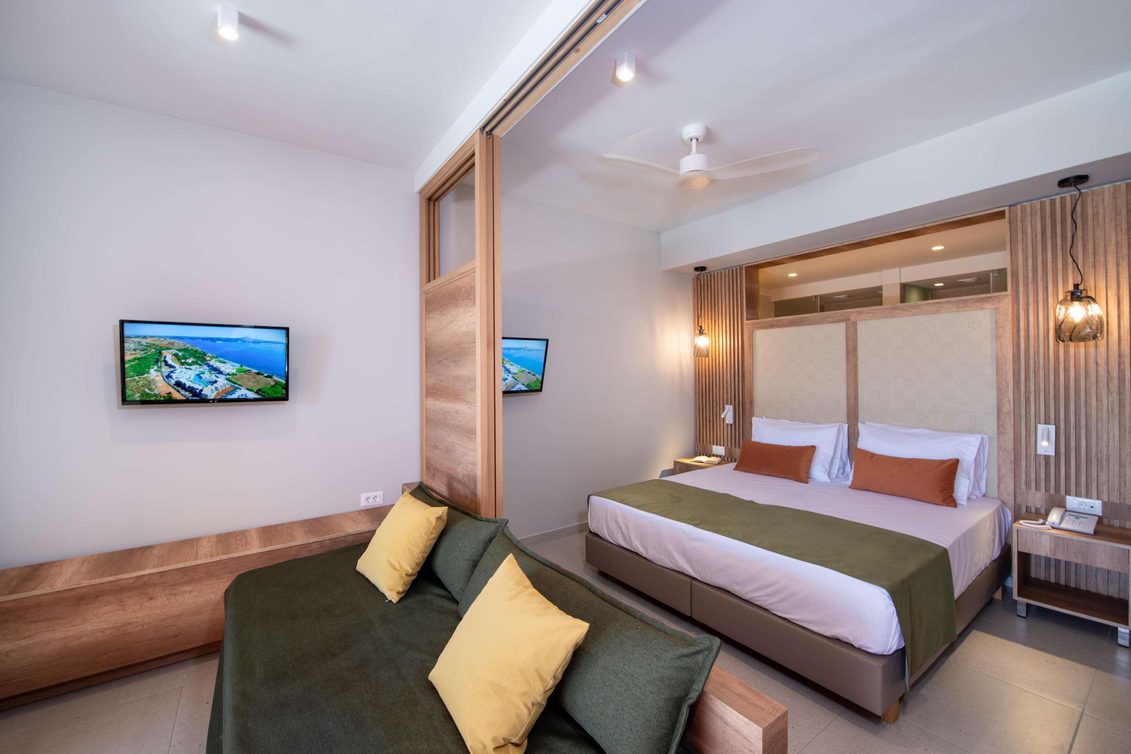A modern hotel room with a large bed, a sofa, a TV, and a ceiling fan.