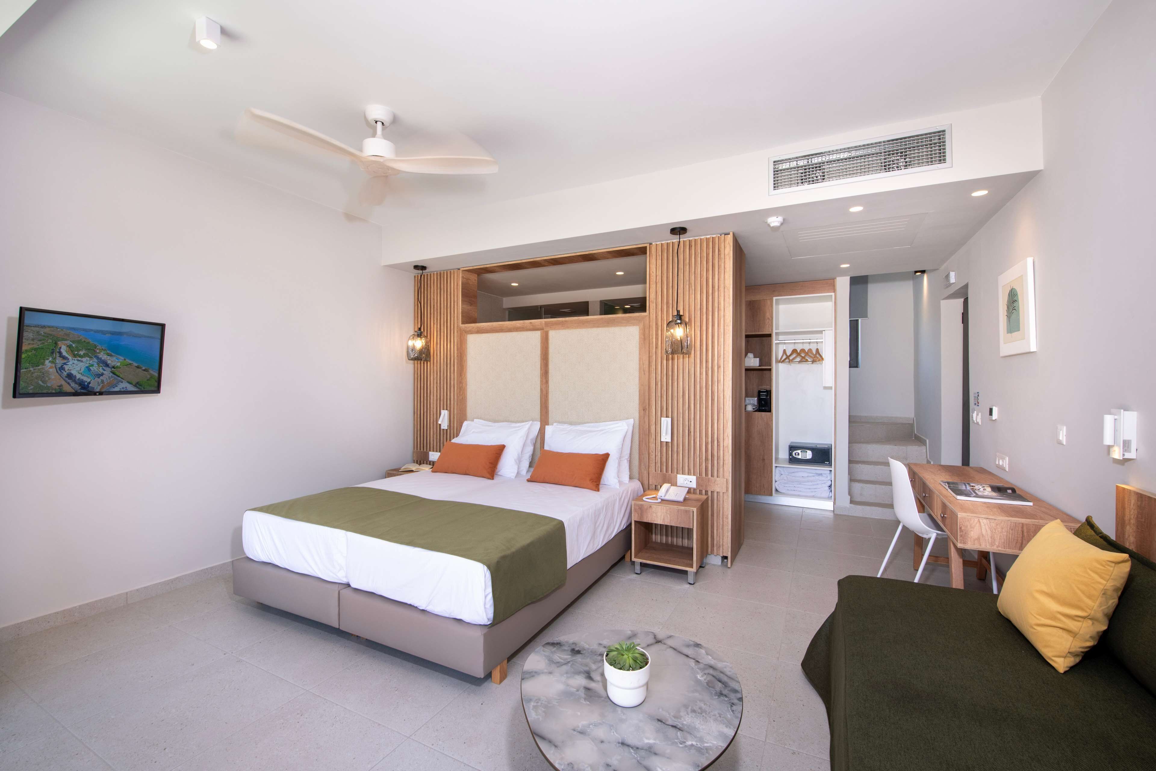 A modern hotel room with a bed, a small table with a plant, a sofa, a desk, and a ceiling fan.