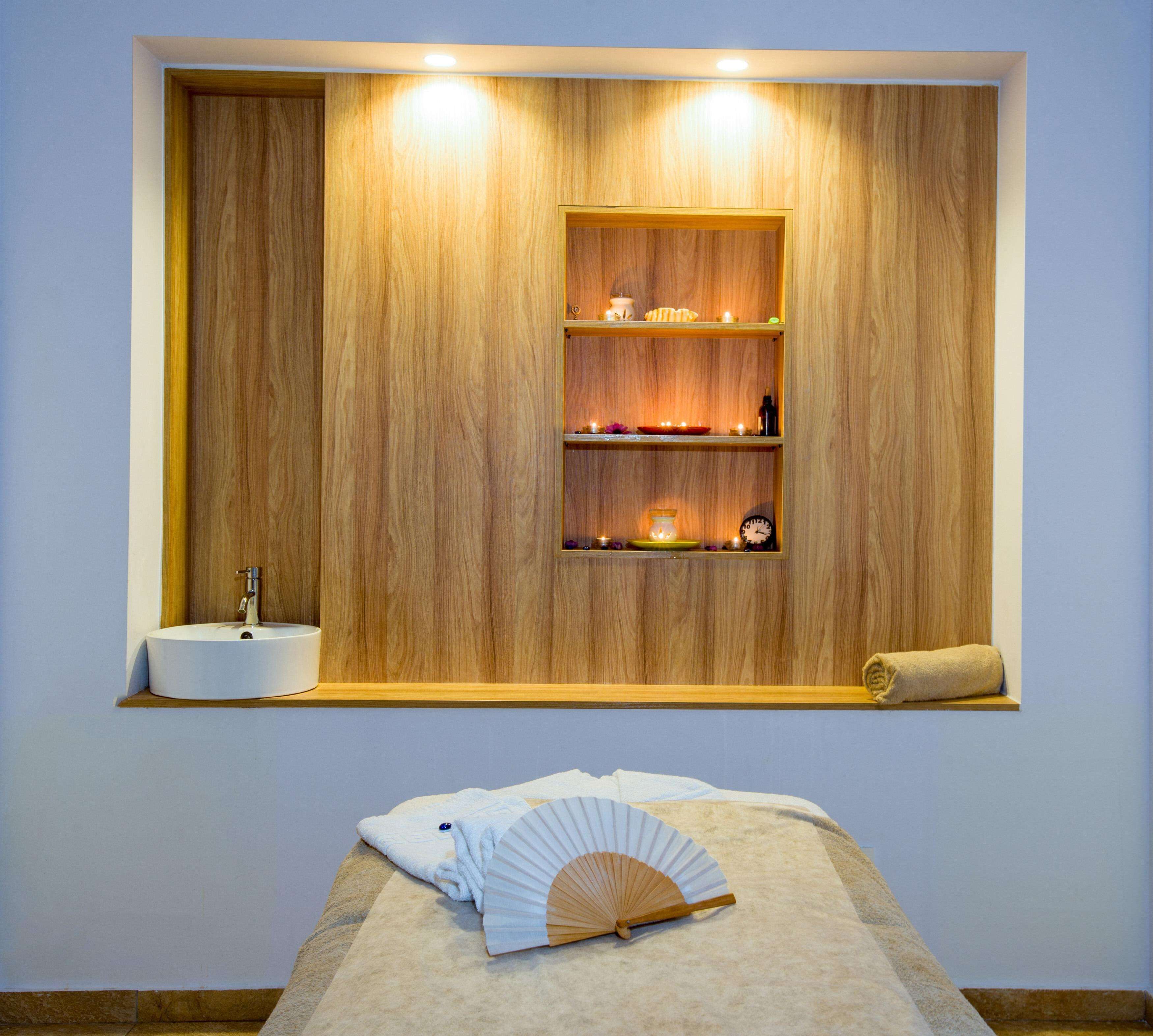 A spa room with a wooden wall, a sink, shelves with candles, and a fan on the floor.