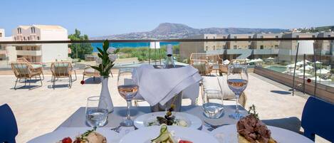 A dining table with three plates of food, a bottle of wine, and glasses on a terrace overlooking a body of water and buildings.