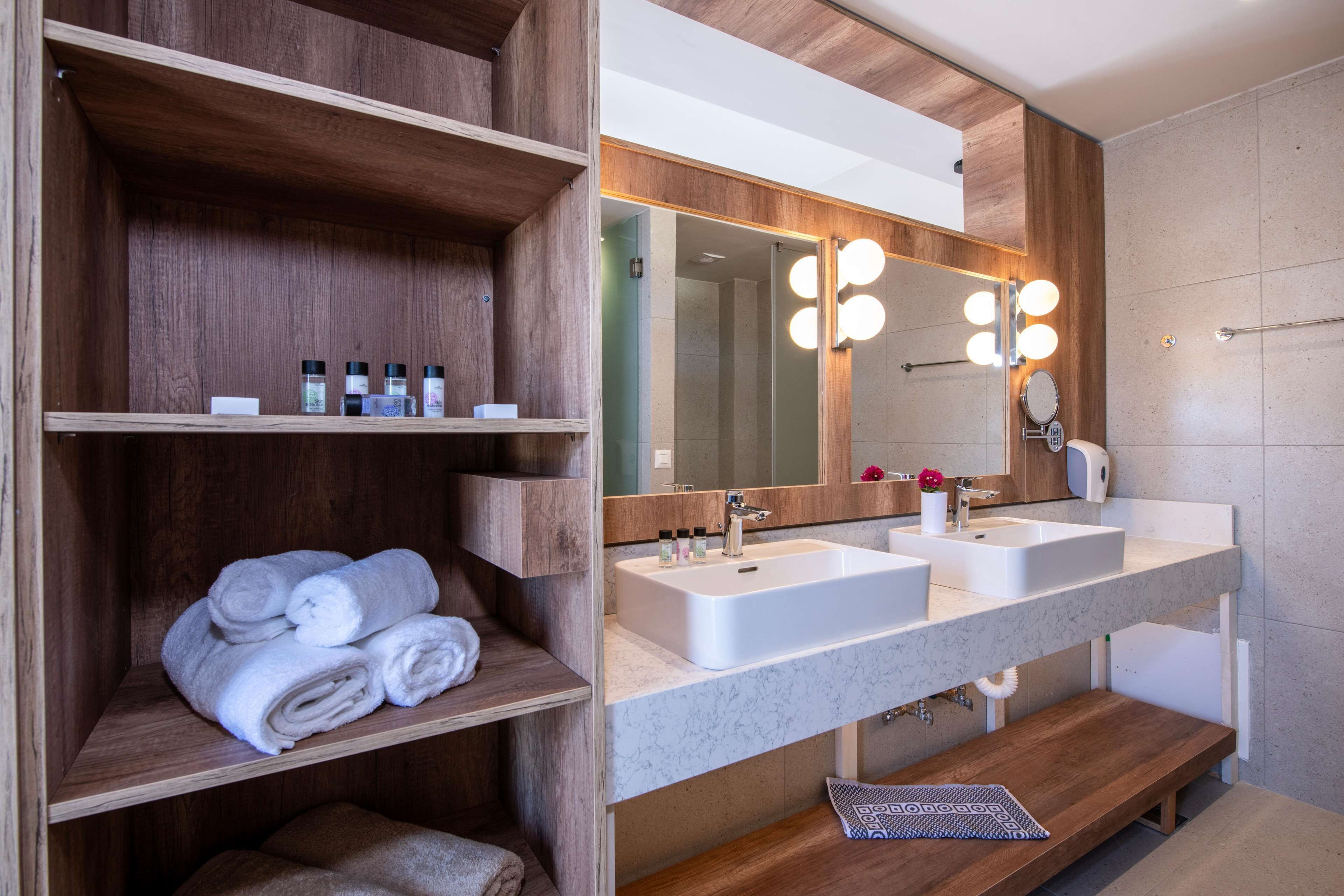 Un baño moderno con estantes de madera, un espejo grande con luces, dos lavabos y un perchero para toallas.