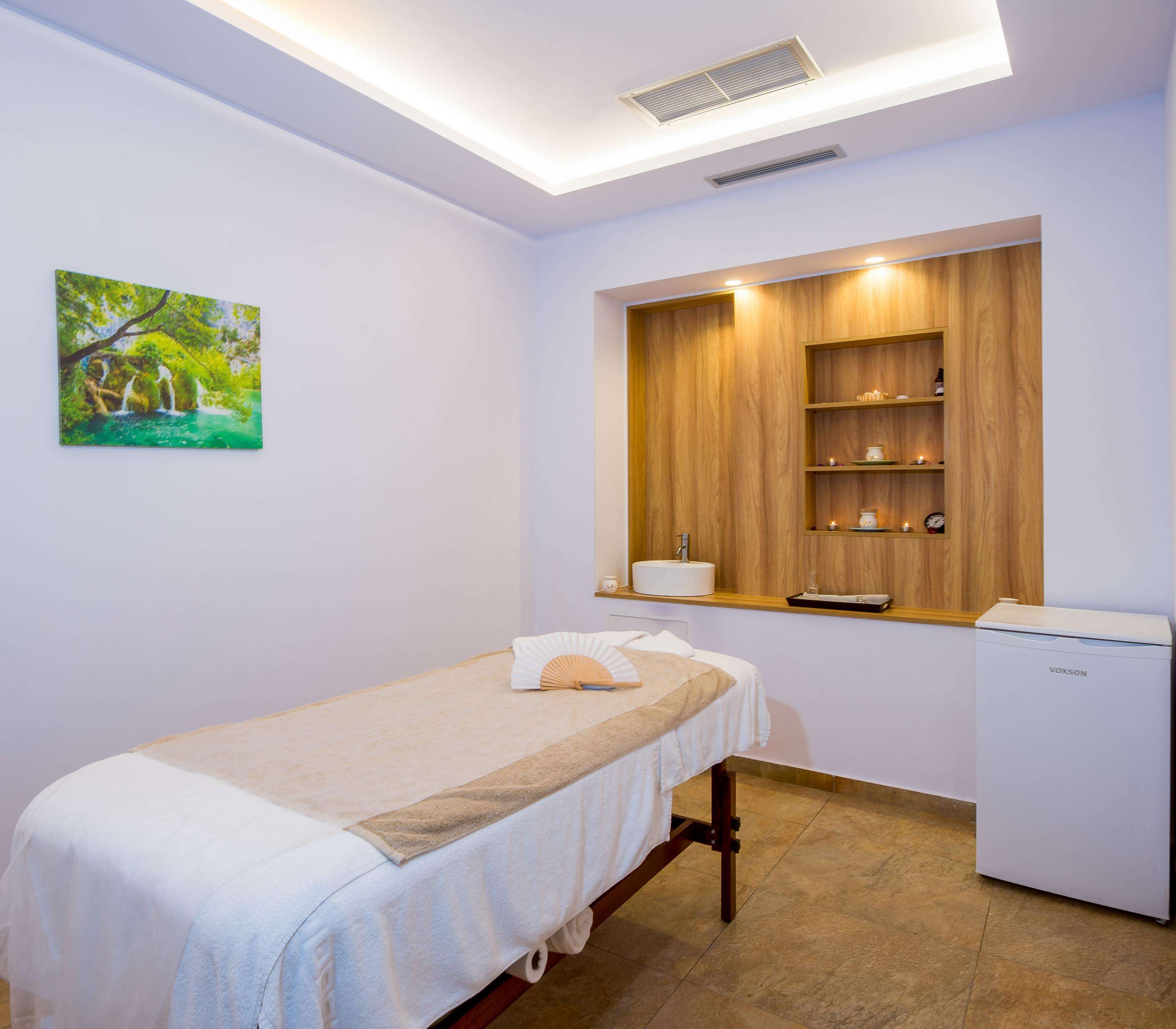Una cama individual con cubierta blanca, una mesita de noche con lámpara, una estantería de madera sobre la cama y un pequeño electrodoméstico blanco a la derecha.