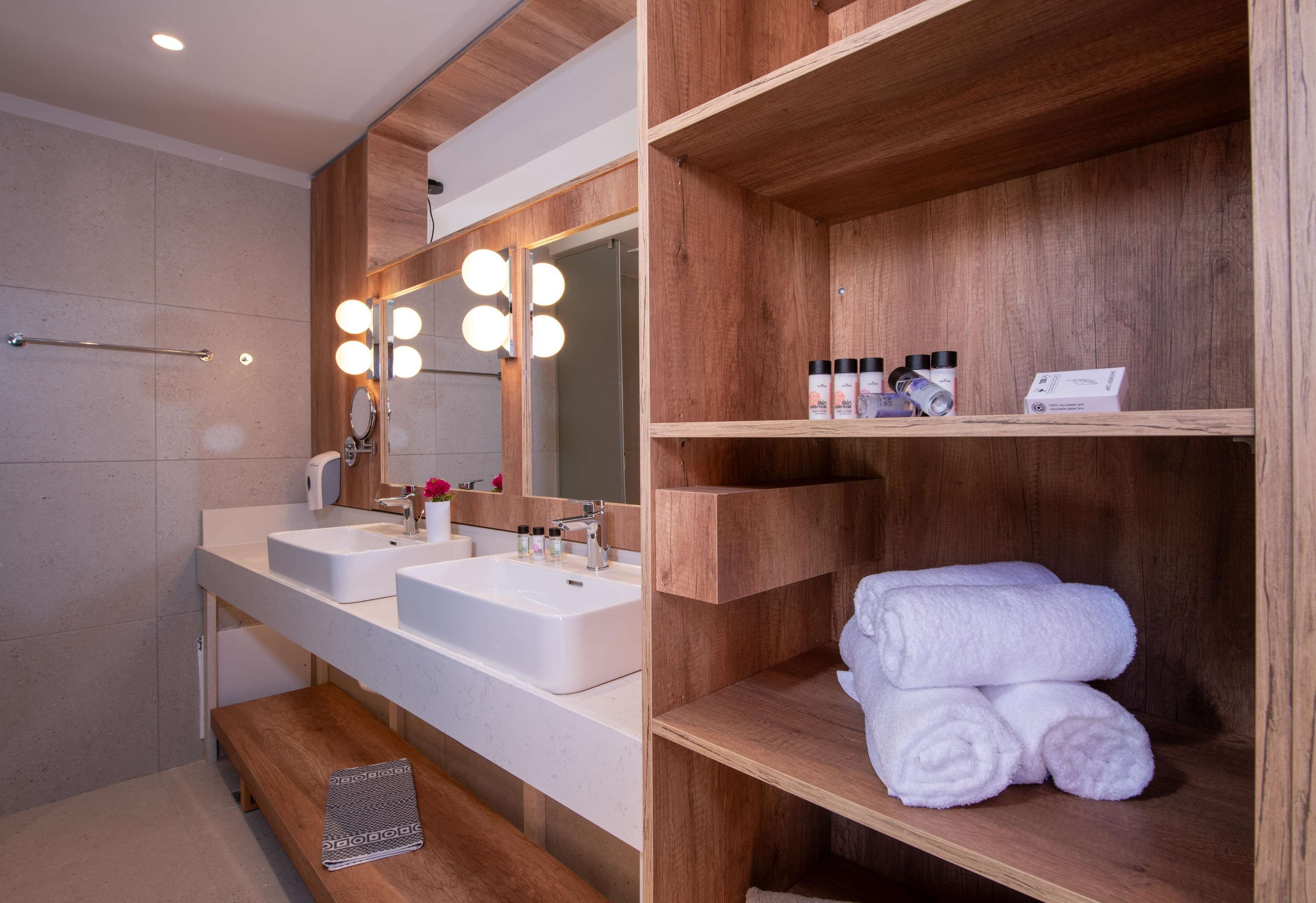 A modern bathroom with two sinks, a large mirror with lights, and wooden shelves with towels.