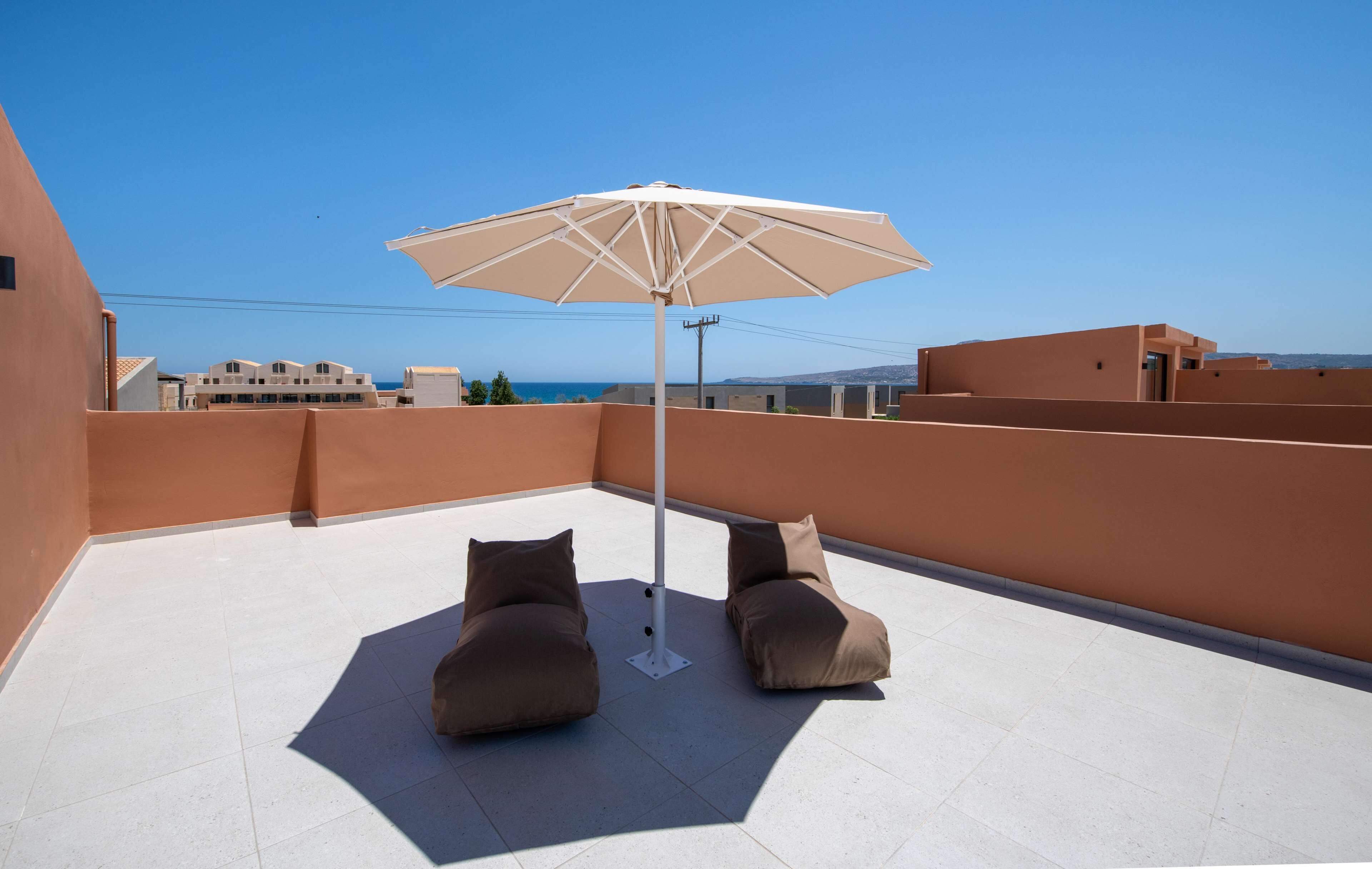 Terraza en la azotea con dos zonas de estar acolchadas y una sombrilla grande, con vistas a un paisaje urbano costero.