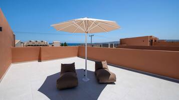 Terraza en la azotea con dos zonas de estar acolchadas y una sombrilla grande, con vistas a un paisaje urbano costero.