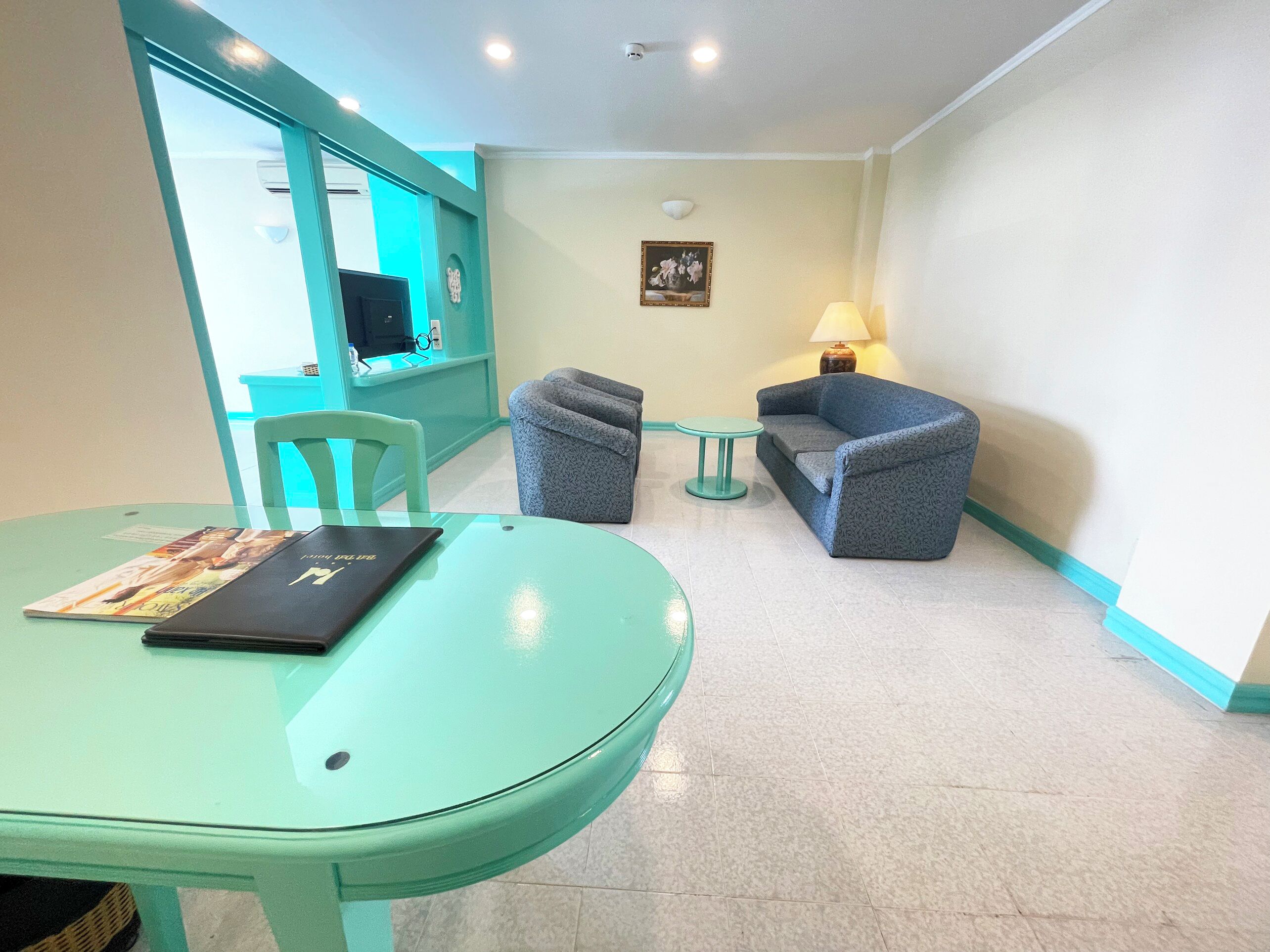 Une pièce avec une table de couleur turquoise, un canapé et une petite table ronde. On y trouve une chaise, une lampe et un tableau accroché au mur.