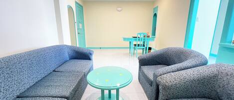 A room with two patterned sofas facing a small turquoise table, a teal dining table with chairs, and a light-colored tiled floor.