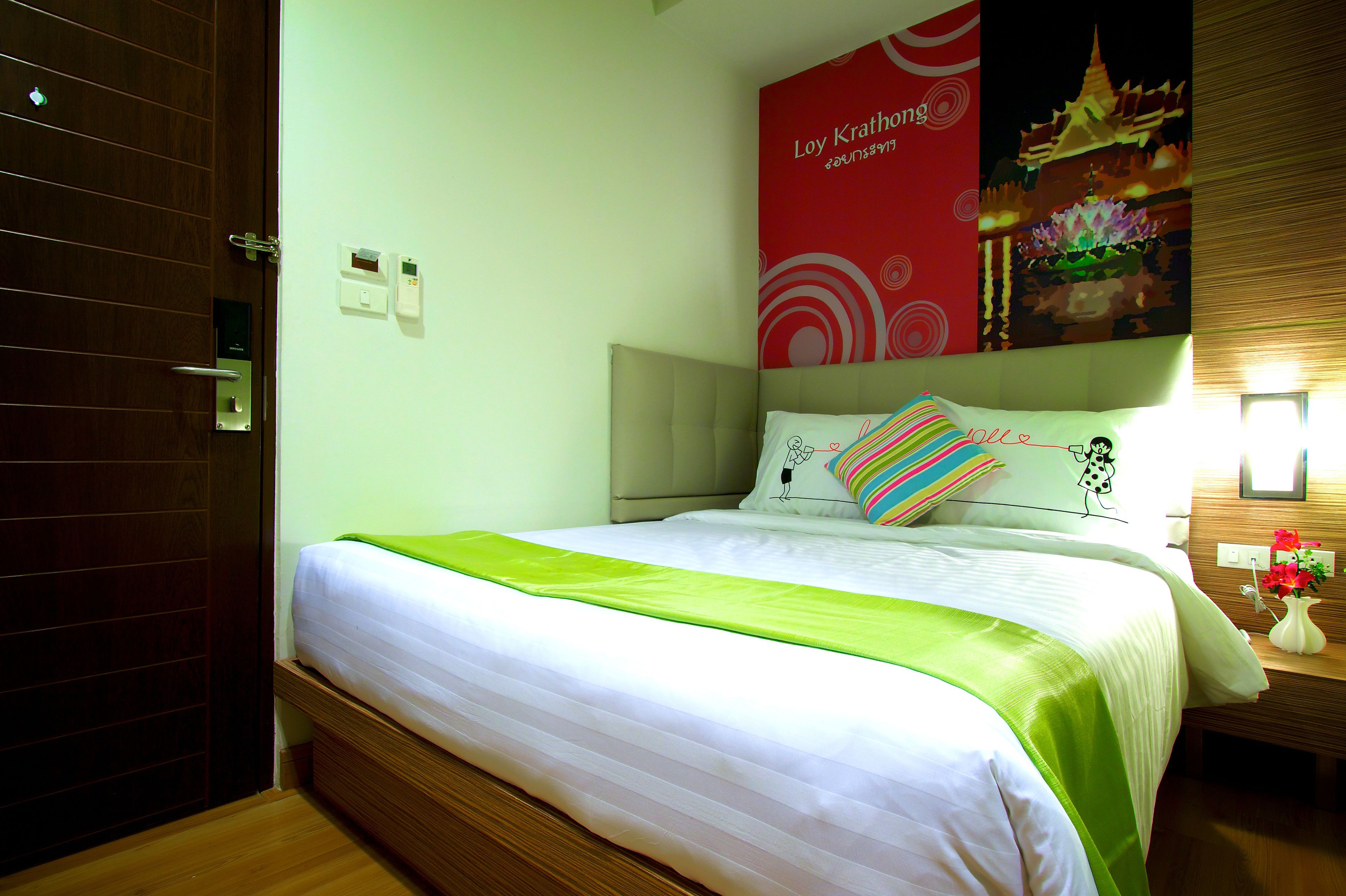 A neatly made bed with a green bedspread, a red wall with a poster, and a wooden nightstand with a vase of flowers.