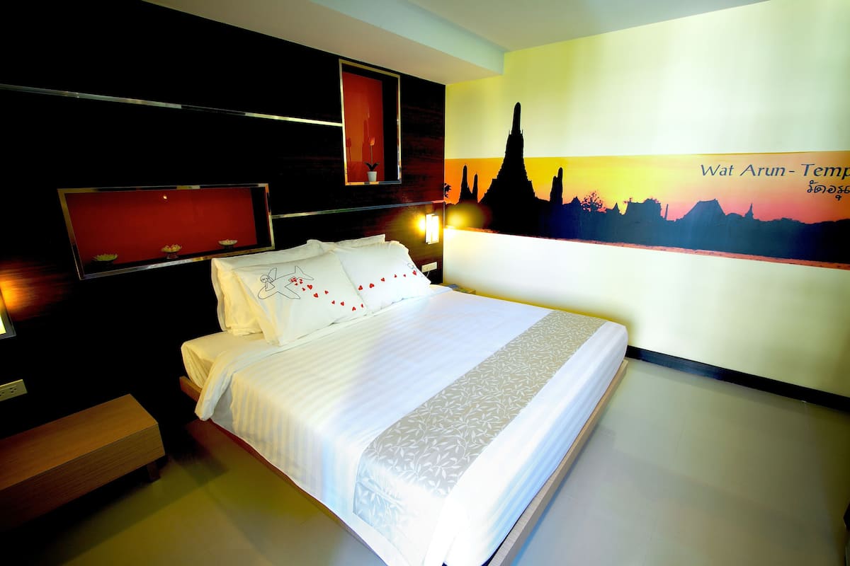 Hotellihuone, jossa on suuri sänky, yöpöytä, seinälle asennettu televisio ja seinämaalaus, jossa on kuvattuna Wat Arun -temppeli.