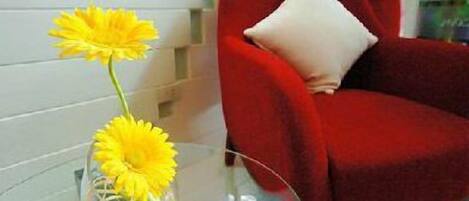 Una mesa de vidrio con un jarrón de flores amarillas, un sillón rojo con un cojín blanco, y una planta al fondo.