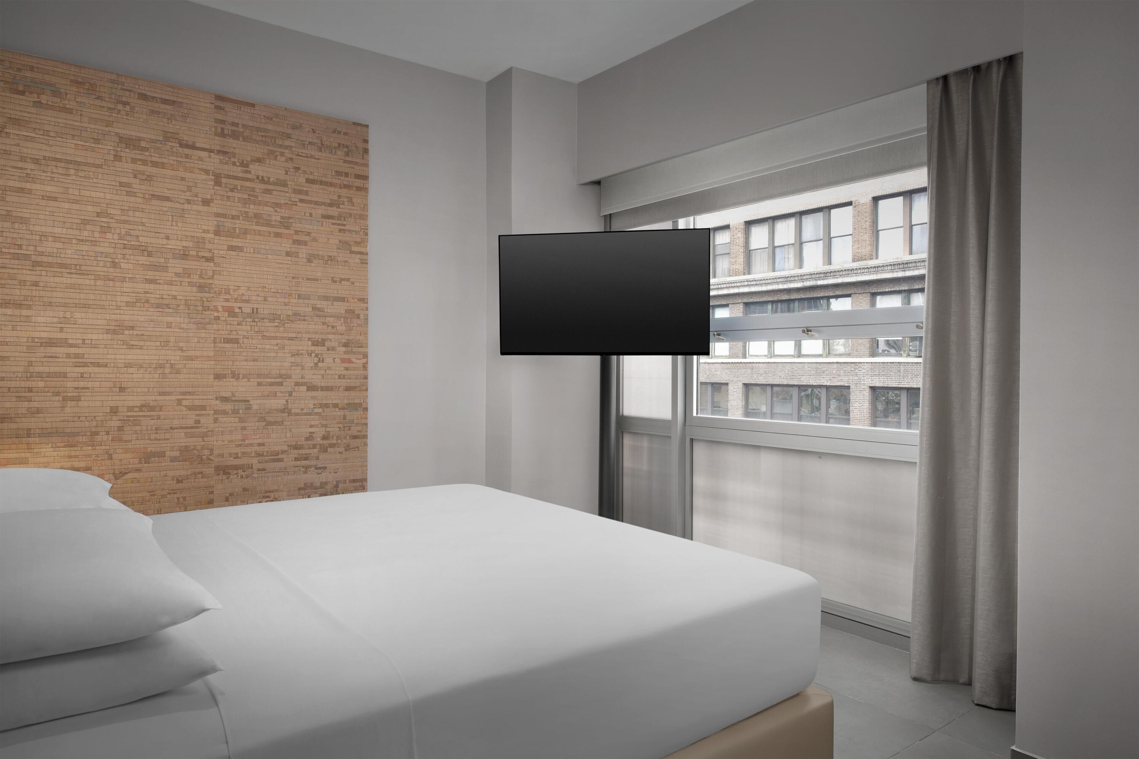 A modern hotel room with a brick accent wall, a large window with curtains, a flat-screen TV, and a neatly made bed.