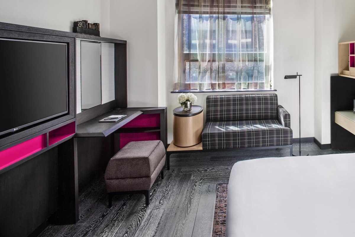 Habitación de hotel con cama, televisión, zona de estar con banco, mesita, lámpara y ventana con cortinas.