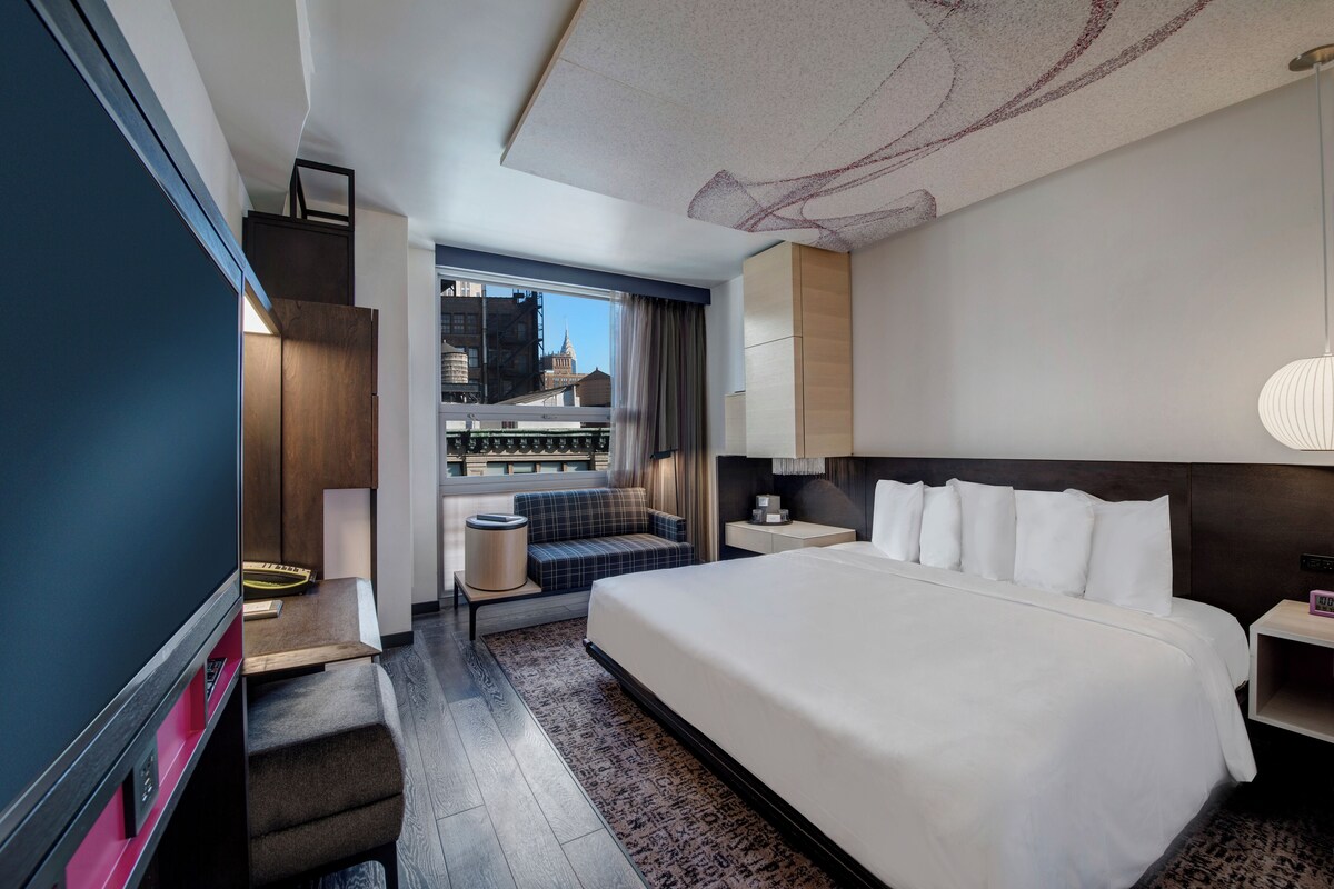 A modern hotel room with a large bed, a desk, a sofa, and a view of the city through the window.