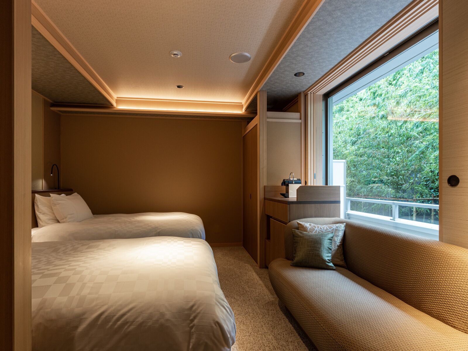 A compact bedroom with two beds, a small seating area, and a window with a view of greenery.