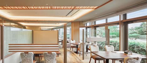 A modern dining area with wooden panel ceilings, large windows offering views of a garden, and a mix of seating arrangements including tables and chairs.