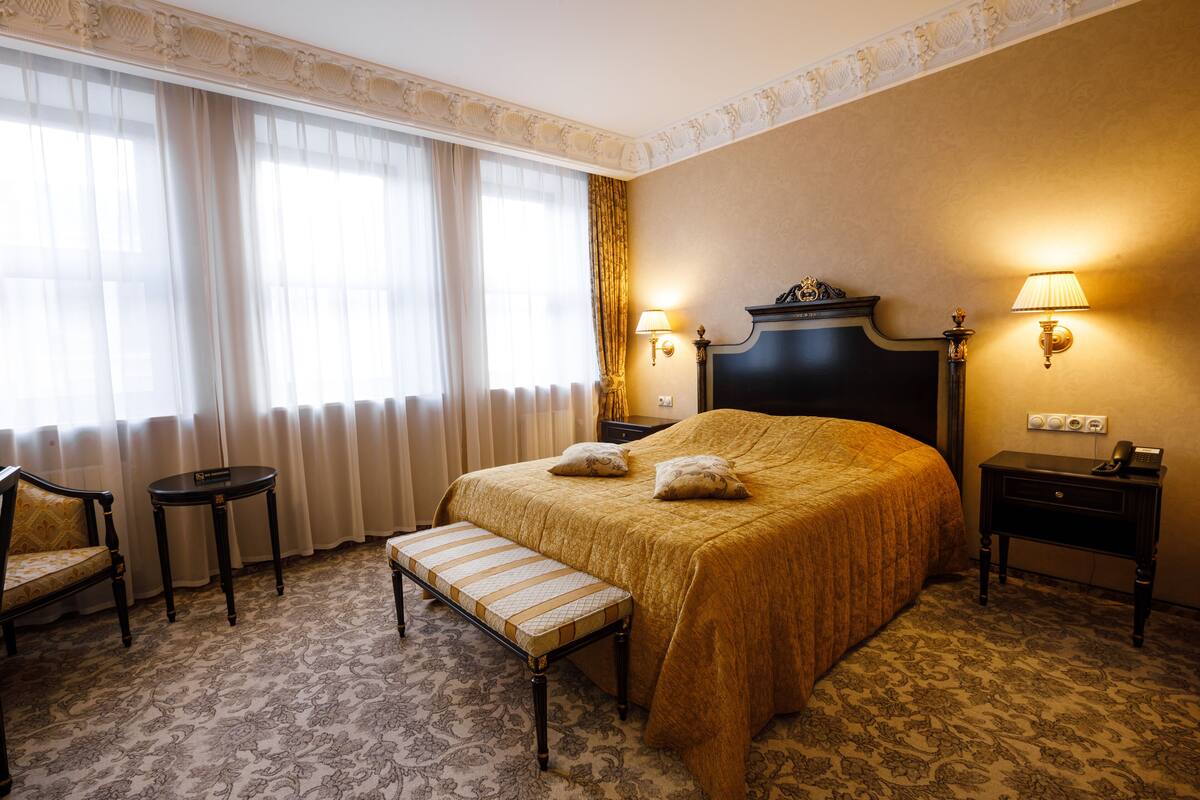Een hotelkamer met een groot bed, twee nachtkastjes, een bankje, een stoel en een raam met gordijnen.