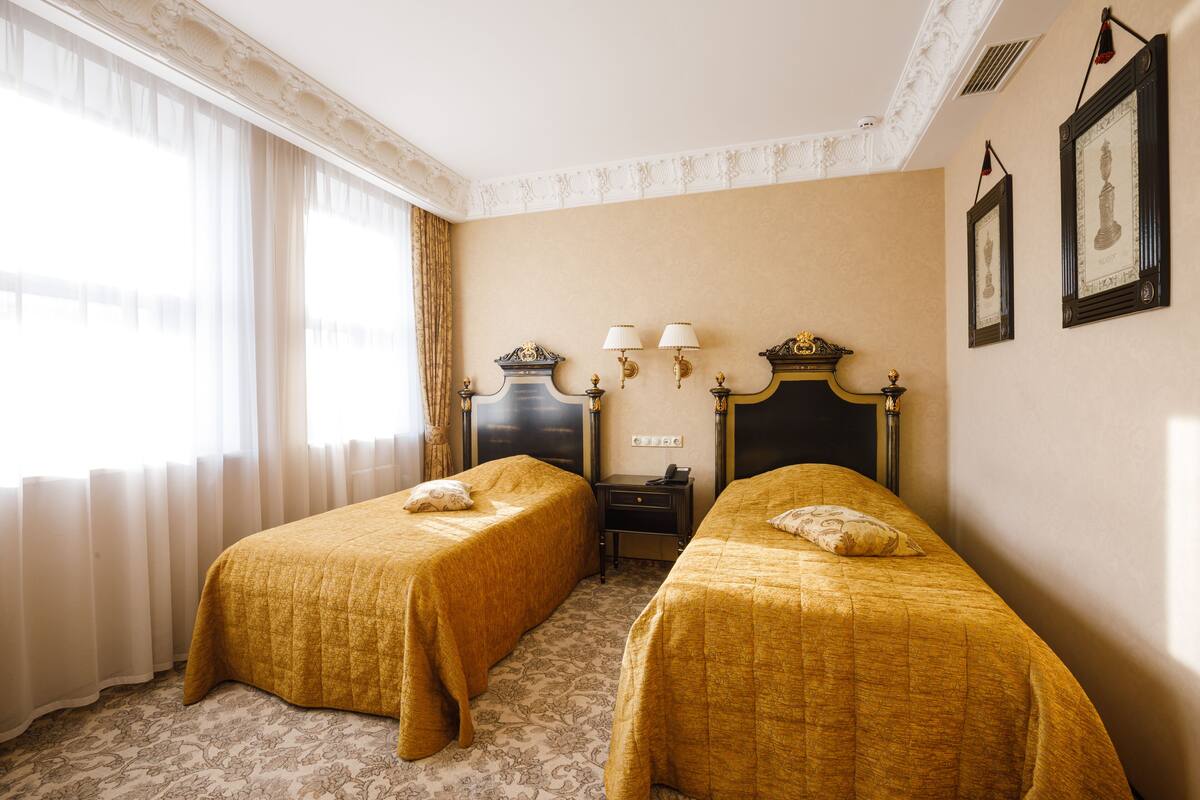 Een hotelkamer met twee eenpersoonsbedden, een nachtkastje, een dressoir en een raam met dunne gordijnen.
