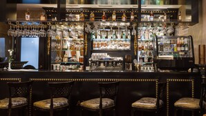 Uma área de bar com várias prateleiras de garrafas de bebidas, copos e uma máquina de café. Há quatro banquetas estofadas.
