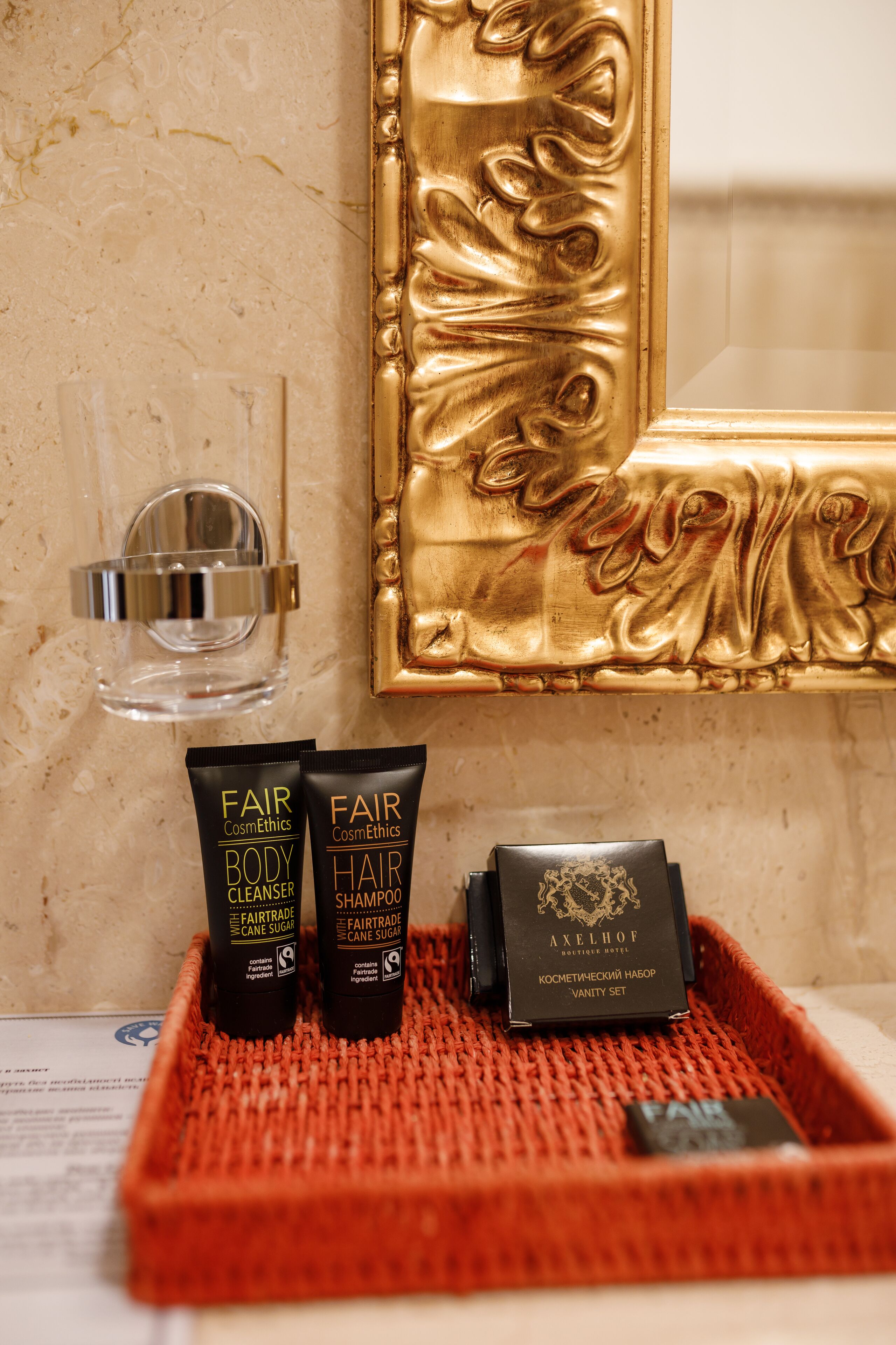 A bathroom vanity with a gold-framed mirror, a red woven tray holding two body cleansers and a hair shampoo, and a small black box labeled "AXELHOF".