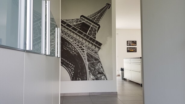 A hallway with a large black and white mural of the Eiffel Tower, white walls, and a white cabinet with a trash bin. - fewosi (Pohlheim)