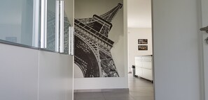 A hallway with a large black and white mural of the Eiffel Tower, white walls, and a white cabinet with a trash bin.