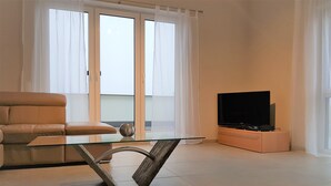 A modern living room with a glass coffee table, a sofa, a flat-screen TV, and a sliding glass door leading to a balcony. - fewosi (Pohlheim)