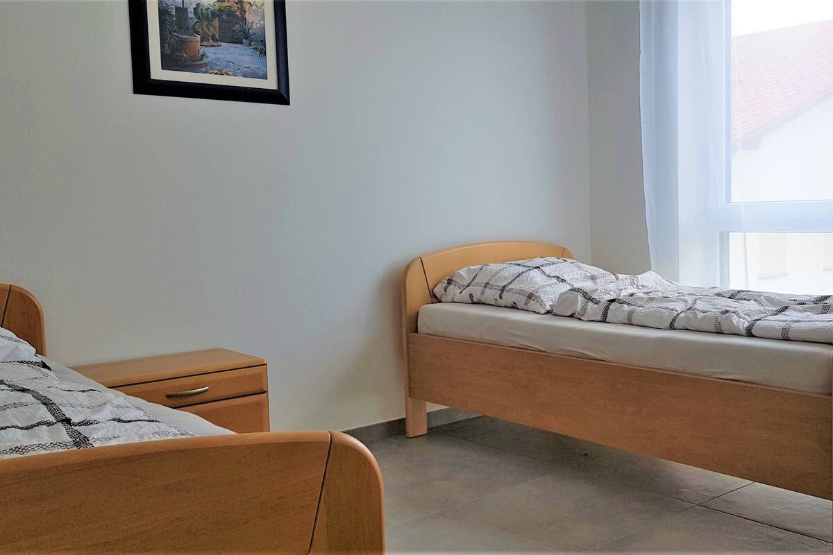 A bedroom with two beds, a nightstand, and a window with a view of a house.