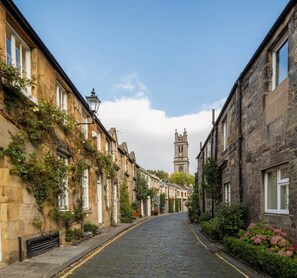 A cobblestone street lined with historic stone buildings, a church tower in the distance, and lush greenery. - St Stephen Street Apartment (Edinburgh)