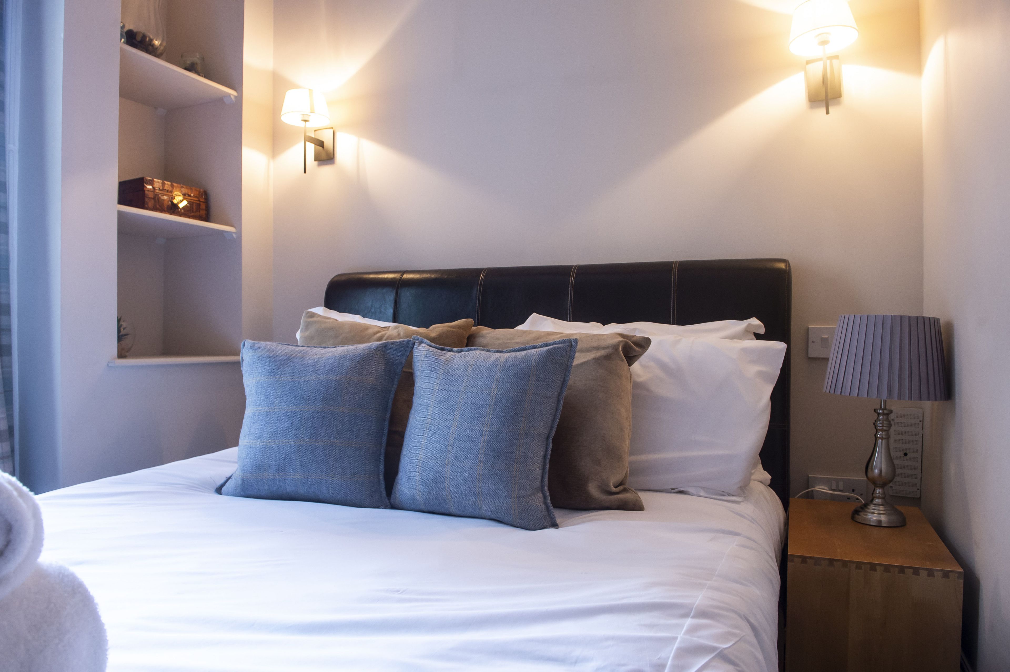 Una cama bien hecha con ropa de cama blanca y gris, un cabecero negro y dos lámparas fijadas a la pared.