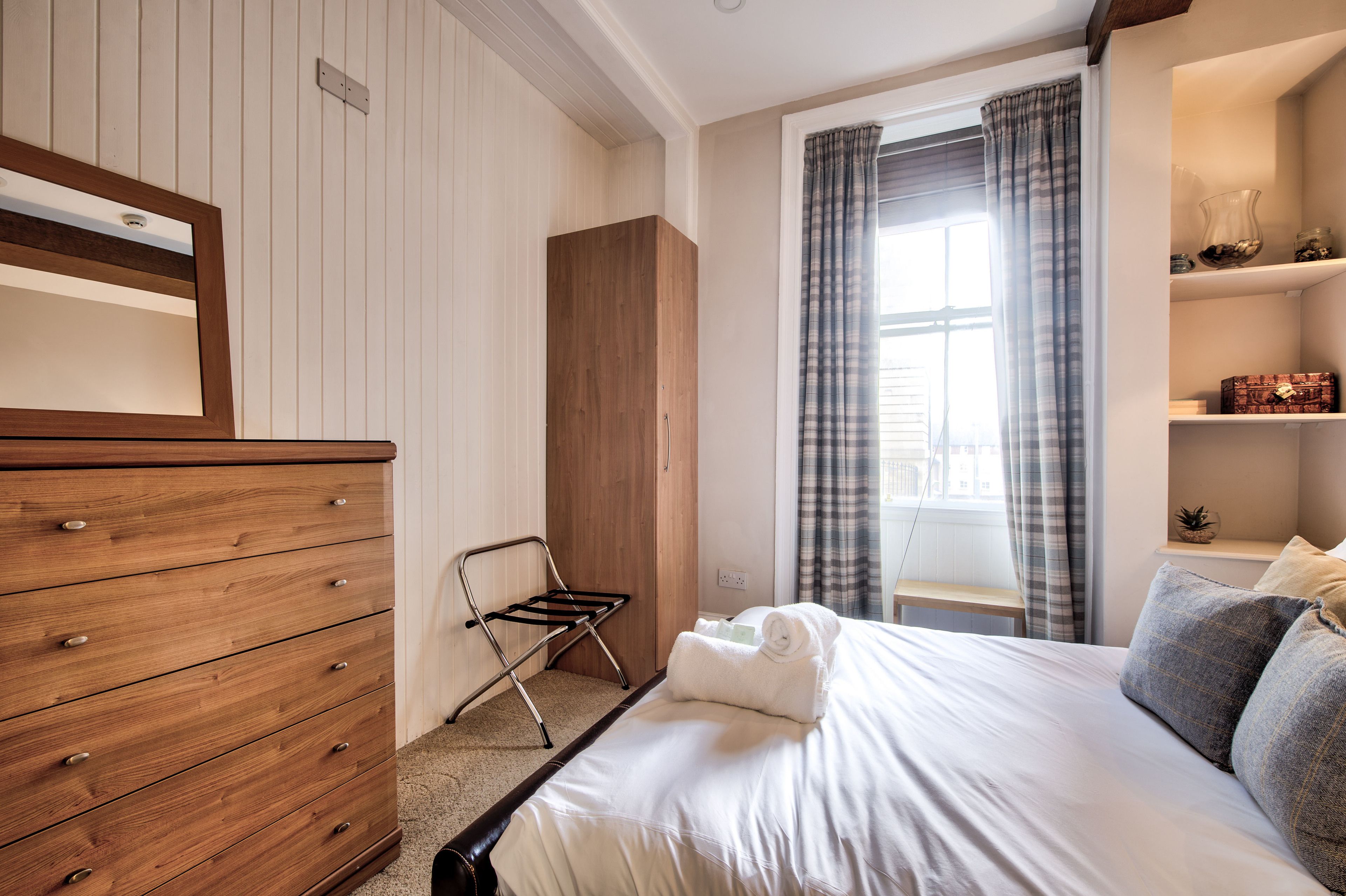 A bedroom with a wooden dresser, a metal chair, a neatly made bed with towels, a window with curtains, and a small shelf with decorative items.