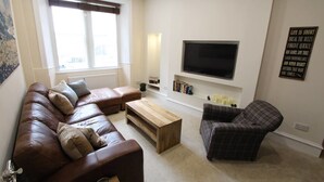 A living room with a brown leather sofa, a plaid armchair, a wooden coffee table, and a flat-screen TV mounted on the wall. - St Stephen Street Apartment (Edinburgh)