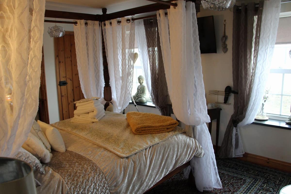 Quarto com cama de dossel, uma janela grande com cortinas, uma estátua de Buda e uma mesa de cabeceira com abajur.