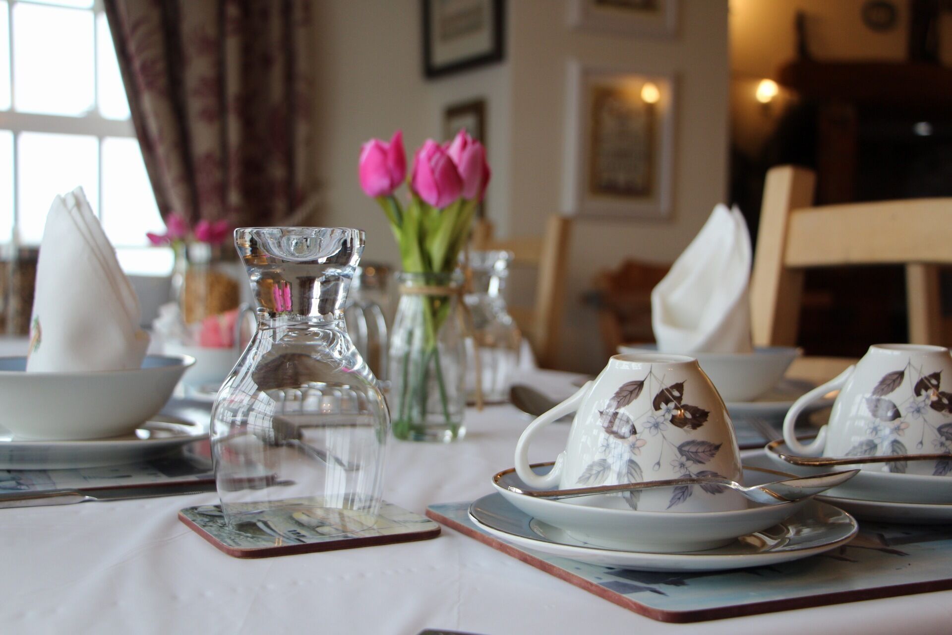 A dining table set with white plates, floral teacups, and a clear glass pitcher.