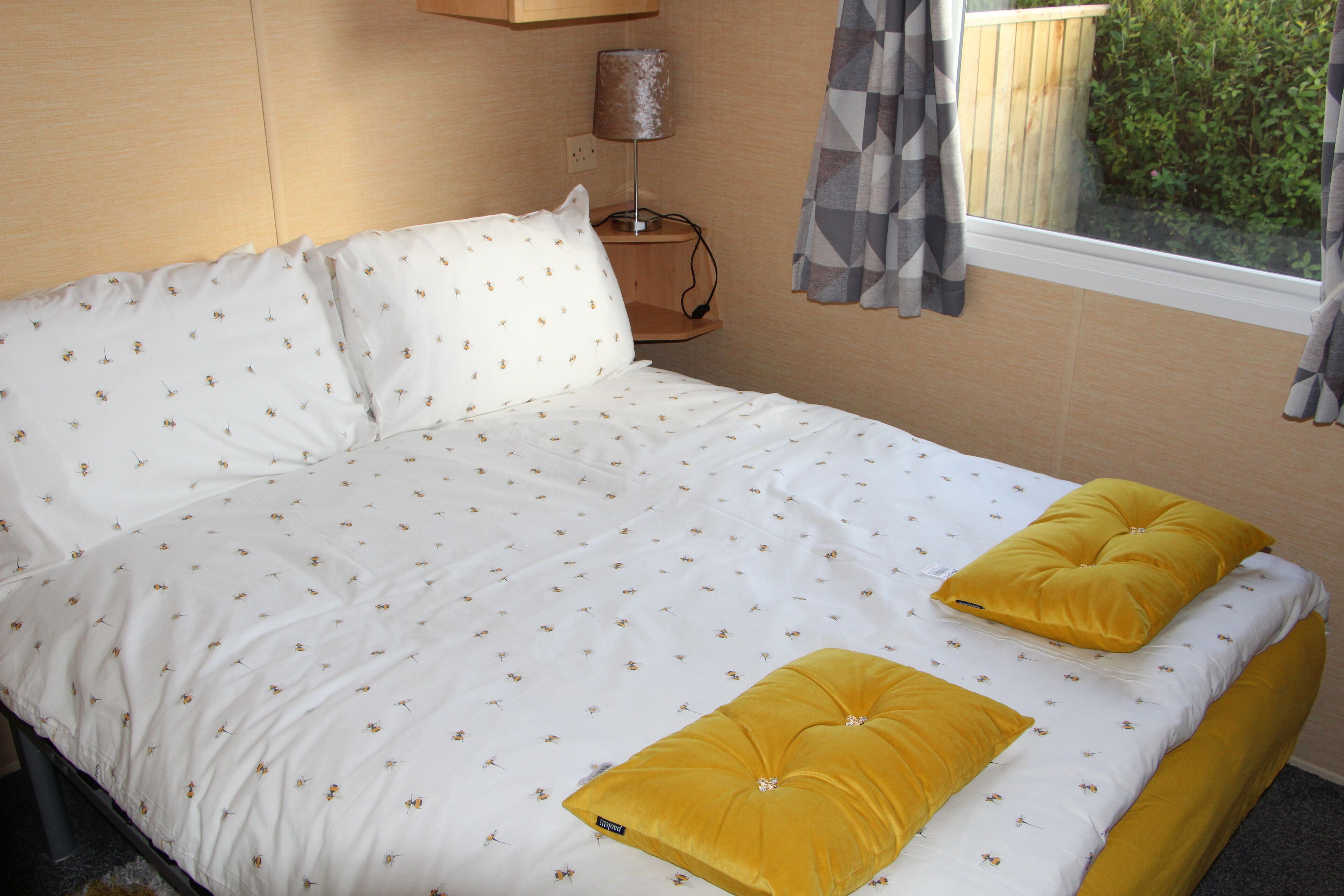 A bed with white bedding and two yellow pillows, a bedside table with a lamp, and a window with curtains.