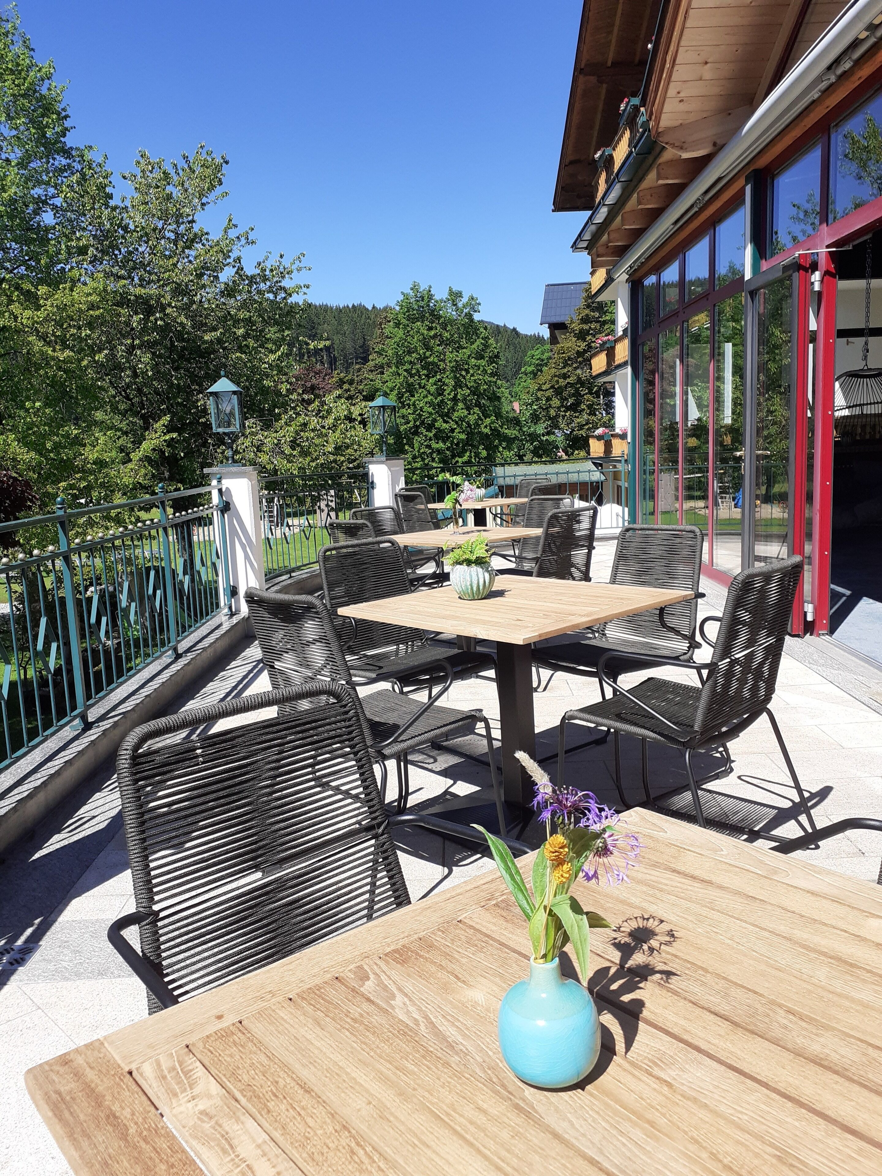 Een terras buiten met een houten tafel en zwarte wickerstoelen, omringd door een metalen hek en glazen deuren die naar een gebouw leiden.