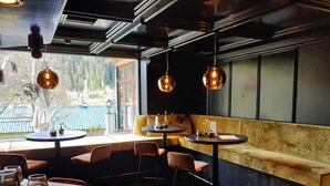 Un restaurante acogedor con mesas redondas y asientos acolchados, ventanas amplias que ofrecen vistas a árboles y una cerca azul, además de lámparas colgantes.