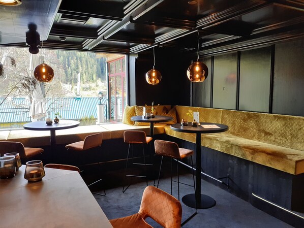 A cozy restaurant with round tables and cushioned seating, large windows offering a view of trees and a blue fence, and pendant lighting. - Hotel Matschner (Ramsau am Dachstein)